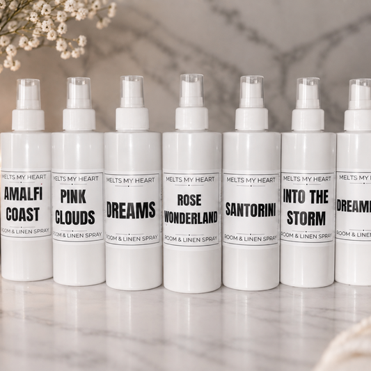 Eight white room and linen spray bottles with clear caps are lined up on a light marble surface. Each bottle has a black label with a different scent name, including Amalfi Coast, Pink Clouds, Dreams, Rose Wonderland, Santorini, Into the Storm, and Dreamland.