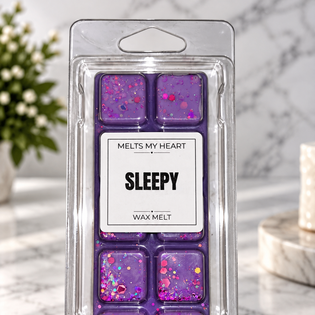 A package of six purple wax melts with pink and silver glitter, labeled 'SLEEPY' by 'MELTS MY HEART', displayed on a marble surface with a blurred background.