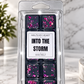 A clear plastic clamshell package containing six dark wax melts with pink and purple glitter, labeled 'MELTS MY HEART INTO THE STORM WAX MELT,' photographed on a light marble surface with a blurred plant and vase in the background.