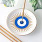 A white ceramic incense holder with a blue and white evil eye design and tan sunburst rays, shown with incense sticks on a light surface with decorative leaves.