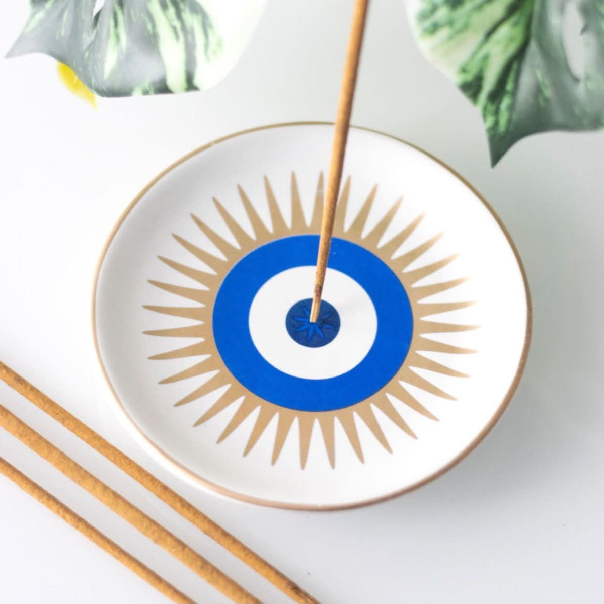 A white ceramic incense holder with a blue and white evil eye design and tan sunburst rays, shown with incense sticks on a light surface with decorative leaves.