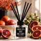 A clear glass reed diffuser bottle with a black cap and black reeds, labeled 'MELTS MY HEART REED DIFFUSER,' styled with blood oranges and sliced citrus around it. Text below reads 'BLOOD ORANGE' with scent notes.
