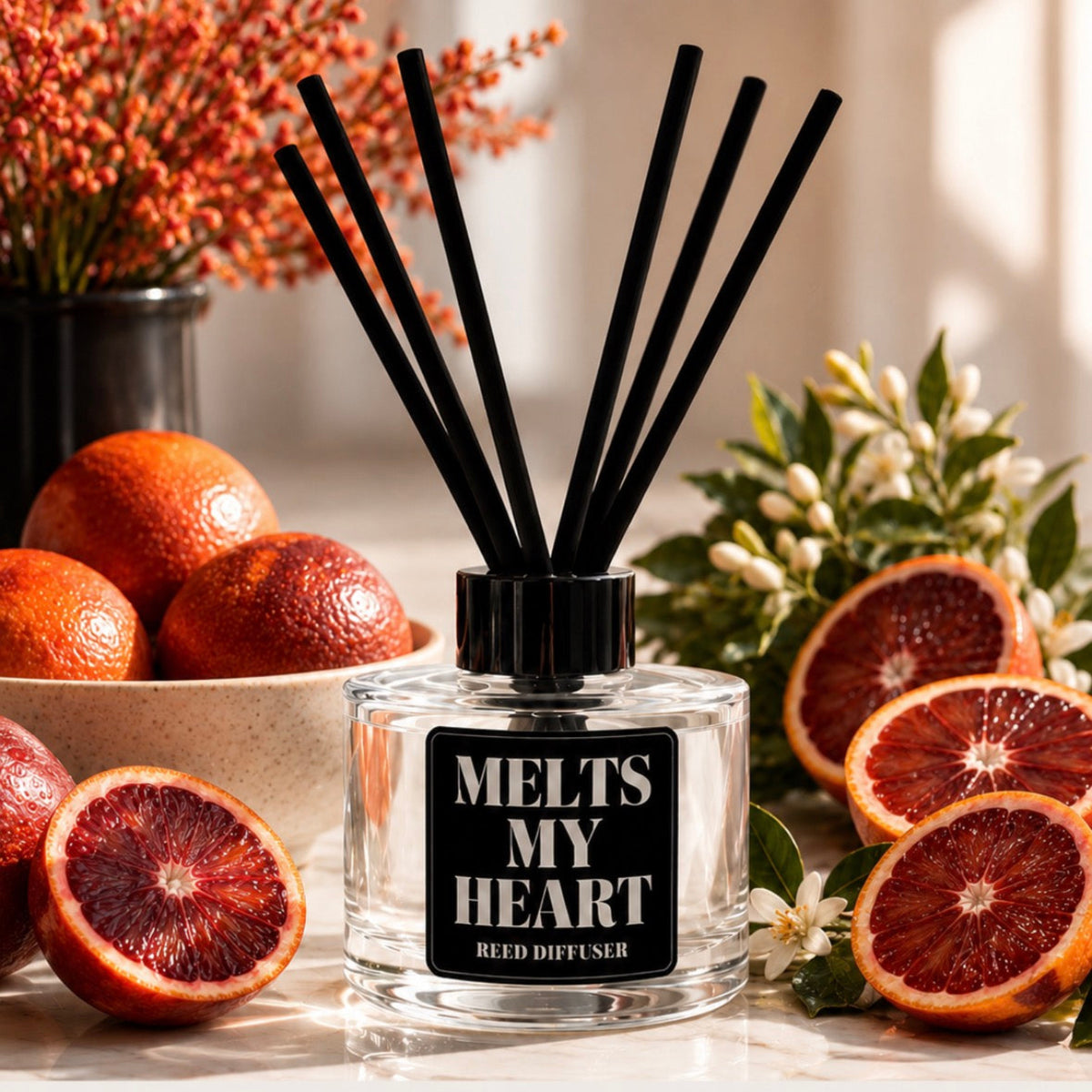 A clear glass reed diffuser bottle with a black cap and black reeds, labeled 'MELTS MY HEART REED DIFFUSER,' styled with blood oranges and sliced citrus around it. Text below reads 'BLOOD ORANGE' with scent notes.