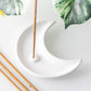 A white crescent moon-shaped incense holder on a light background, with a incense stick inserted and additional sticks placed nearby, with green leaves in the background.
