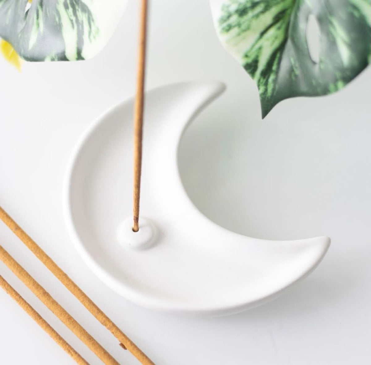 A white crescent moon-shaped incense holder on a light background, with a incense stick inserted and additional sticks placed nearby, with green leaves in the background.