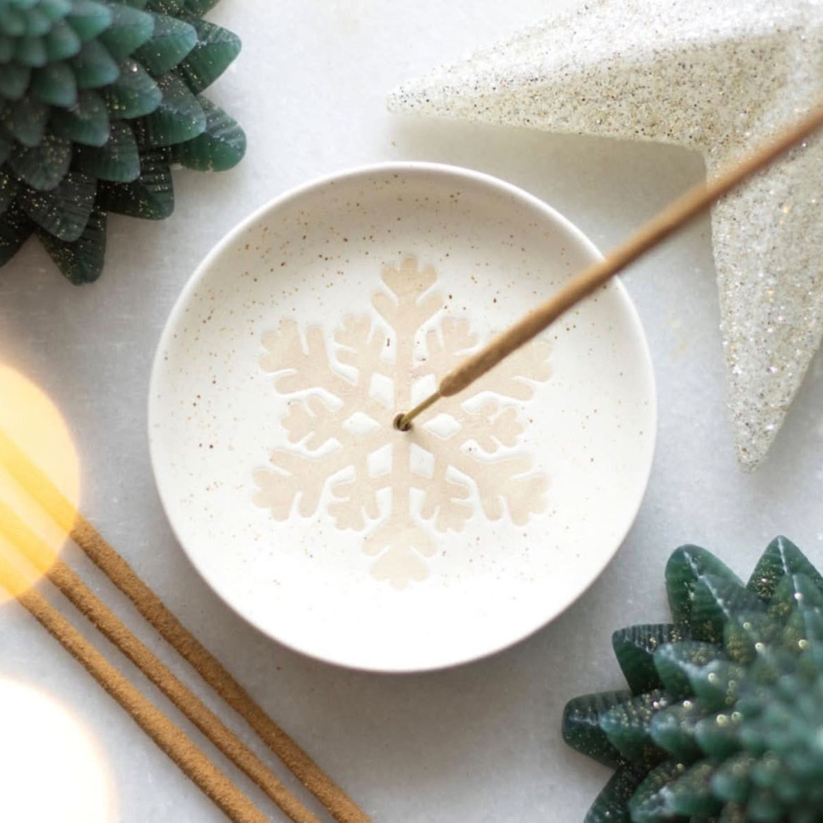 A round white ceramic dish with a subtle speckled texture and a printed snowflake design in the center, shown with wooden incense sticks. The dish is styled with green succulent plants, a glittery star ornament, and warm bokeh light effects on a light surface.